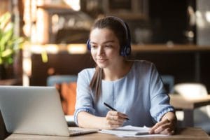 A woman wearing a headset, working on a computer, and taking notes simultaneously